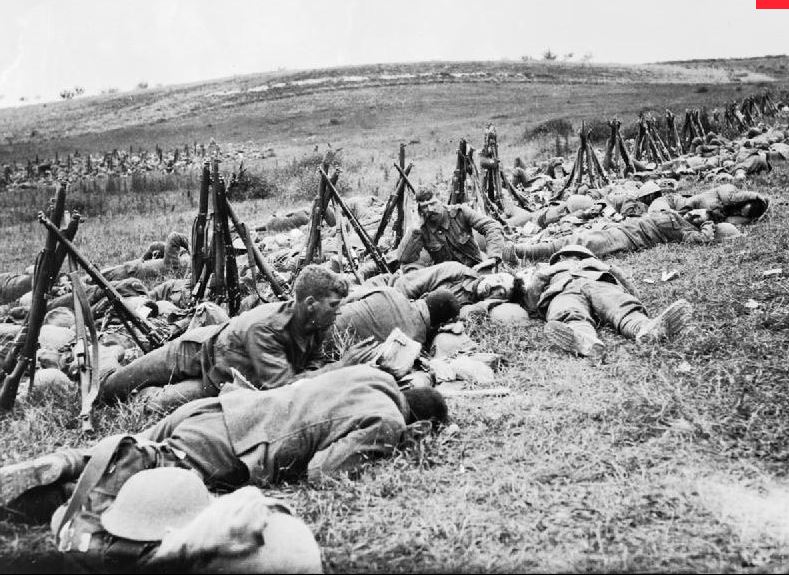 Men of the Royal Warwickshire Regiment, their rifles stacked nearby, lying exhausted in the grass in a rear area.